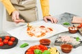 Hands of a caucasian teenage girl in an apron smear tomato sauce on a heart-shaped pizza dough. Royalty Free Stock Photo