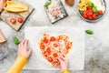 Hands of caucasian teenage girl lay down sliced cherry tomato on heart-shaped pizza dough. Royalty Free Stock Photo