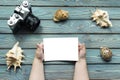 Hands of a child hold a clean white photo mock up, a camera, sea shells, a blue wooden background.