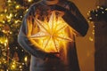 Hands in cozy sweater holding golden illuminated christmas star on background of stylish decorated christmas tree with golden