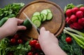 Hands cutting cucumber with a white ceramic knife on a round wooden chopping board. Around the board are ripe fresh vegetables: Royalty Free Stock Photo