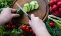 Hands cutting cucumbers with a white ceramic knife on a round wooden chopping board. Around the board are ripe fresh vegetables: Royalty Free Stock Photo