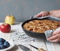 Hands of an elderly woman serve a frying pan with a ready made pie with apples and plums on a wooden white table. Homemade cakes.