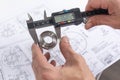 Hands of an engineer measures a metal part with a digital vernier caliper against the background of technical drawings. Quality