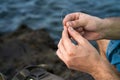 The hands of a fisherman taking the line to tie a knot, in the background a backpack on some rocks and sea water