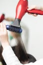 Hands of hairstylist drying brunette hair of client using red hairdryer and blue comb in professional beauty salon