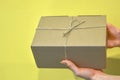 Hands hold a cardboard box against a yellow wall. Gift or packaging made of kraft paper, tied criss-cross with jute rope, on a yel