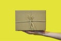 Hands hold a cardboard box against a yellow wall. Gift or packaging made of kraft paper, tied criss-cross with jute rope, on a yel
