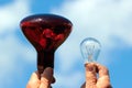 Hands hold infrared lamp and transparent incandescent light bulb against blue sky background Royalty Free Stock Photo