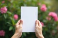 Hands holding blank card on green leaves, pink flowers. Paper sheet template for message, showing information, presentation.