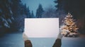 Hands holding a blank white paper against snowy landscape with christmas tree