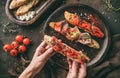 Hands holding a bruschetta with prosciutto, tomatoes, parmesan cheese, sauce pesto, grilled pepper on ciabatta bread on wooden