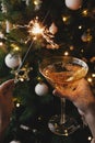 Hands holding burning sparkler and champagne glass against christmas tree lights. Happy New Year Royalty Free Stock Photo