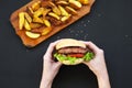 Hands holding fresh burger. Fried potatoes on wooden board. Royalty Free Stock Photo