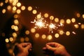 Hands holding glowing sparklers in on background of golden christmas lights, couple celebrating in dark festive room. Happy New Royalty Free Stock Photo