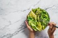 Hands holding healthy fresh green salad with avocado, kiwi, apple, cucumber, pear, greens and sesame on light marble background. Royalty Free Stock Photo