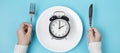 Hands holding knife and fork above alarm clock on white plate on blue background. Intermittent fasting, Ketogenic dieting, weight Royalty Free Stock Photo