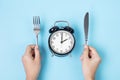 Hands holding knife and fork above alarm clock on white plate on blue background. Intermittent fasting, Ketogenic dieting, weight Royalty Free Stock Photo