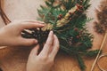 Hands holding pine cones, berries, fir branches, thread, scissors on wooden table. Authentic rural wreath. Christmas wreath