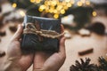 Hands holding rustic christmas gift in black wrapping paper with cinnamon decoration on background of golden lights, pine branches Royalty Free Stock Photo