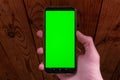 Hands holding smartphone with green display upright on dark wooden background. Moke up