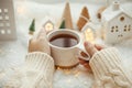 Hands holding stylish cup of tea with modern cute christmas houses, pine cone, wooden star and tree, golden lights on soft warm Royalty Free Stock Photo