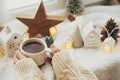 Hands holding stylish cup of tea with modern cute christmas houses, pine cone, wooden star and tree, golden lights on soft warm Royalty Free Stock Photo