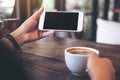 Hands holding white mobile phone with blank black desktop screen and a coffee cup on wooden table in vintage cafe Royalty Free Stock Photo