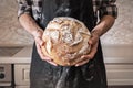 Hands of man holding big loaf of white bread. Male in black apron in home kitchen background with wheat bread