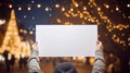 Hands of a man holding a blank sheet of paper in front of a Christmas tree