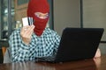 Hands of masked hacker wearing a balaclava holding credit card between stealing data from laptop. Internet crime concept.