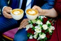 Hands of the newlyweds are holding cups of coffee, a bouquet in the background