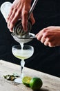 Hands pour through a sieve into a glass cocktail Daiquiri on a wooden table on black background