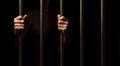 Hands of a prisoner behind prison bars on black background