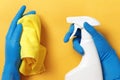 Hands in protective rubber gloves holding a spray bottle with detergent and a cloth on a yellow background. Hygiene prevention.