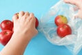 Hands put red apples in polythene bag on blue background, zero waste concept