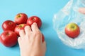 Hands put red apples in polythene bag on blue background, zero waste concept