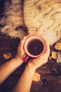 Hands in red knitted gloves holding a hot cup of coffee against yellow leaves background. Concept of autumn time, warmth and cozin