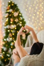Hands in the shape of a heart on the background of the Christmas tree. Girl on sofa cozy heart background. Hygge style with yello