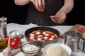 Hands of the woman cook sprinkle Italian raw pizza margarita oreano on a dark background. On the white table lie the ingredients o