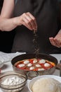 Hands of the woman cook sprinkle Italian raw pizza margarita oreano on a dark background. On the white table lie the ingredients o