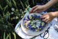 Hands of woman is making wreath of cornflowers and chamomile on white tablecloth in wheat field background