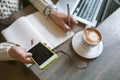 Hands of woman with pen and notepad, holding smartphone with coffee