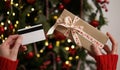 Hands of a woman in a red sweater holding a credit card and a gift box on the background of a Christmas tree close-up. Christmas Royalty Free Stock Photo