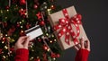 Hands of a woman in a red sweater holding a credit card and a gift box on the background of a Christmas tree close-up. Christmas Royalty Free Stock Photo