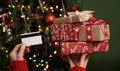 Hands of a woman in a red sweater holding a credit card and a gift box on the background of a Christmas tree close-up. Christmas Royalty Free Stock Photo