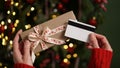 Hands of a woman in a red sweater holding a credit card and a gift box on the background of a Christmas tree close-up. Christmas Royalty Free Stock Photo