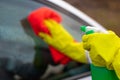 Hands in yellow rubber gloves wipe the glass of a car with a red rag and spray water from a spray gun on a warm autumn day Royalty Free Stock Photo