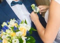 The hands of the young bride correct the boutonniere on the blue suit of the groom with a wedding bouquet in his hands