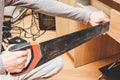 The hand of a young caucasian guy is sawing a board with a saw from the back of a desk in his room.